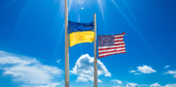 Bright blue sky with a few white clouds in the background. The Ukraine flag waves from a silver flag pole with an American flag waving from another silver flag pole next to it.