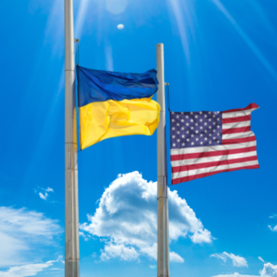 Bright blue sky with a few white clouds in the background. The Ukraine flag waves from a silver flag pole with an American flag waving from another silver flag pole next to it.